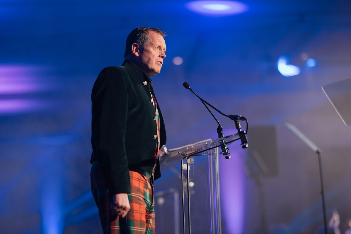 A professional speaker wearing a velvet jacket and tartan trousers delivers a talk at a formal corporate event in Edinburgh, photographed under blue and purple stage lighting.
